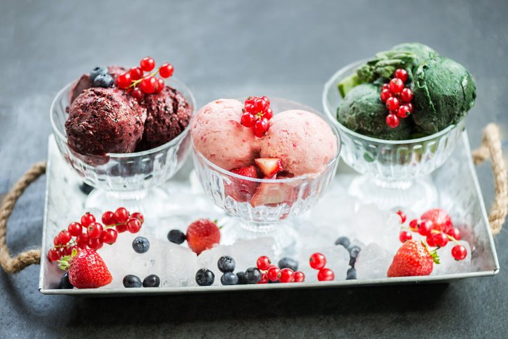 trzy, szklane pucharki, na metalowej tacy wypełnione wegańskimi lodami, w otoczeniu świeżych owoców; borówek, truskawek i czerwonych porzeczek